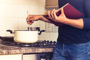 A man reading a cookbook and stirring a pot on the stove.