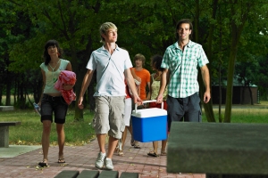 A group of people with two young men carrying a cooler and walking towards a picnic table.