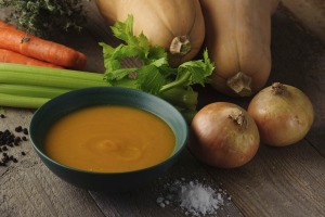 A bowl of butternut squash soup with whole celery, carrots, onions, and butternut squash next to it.